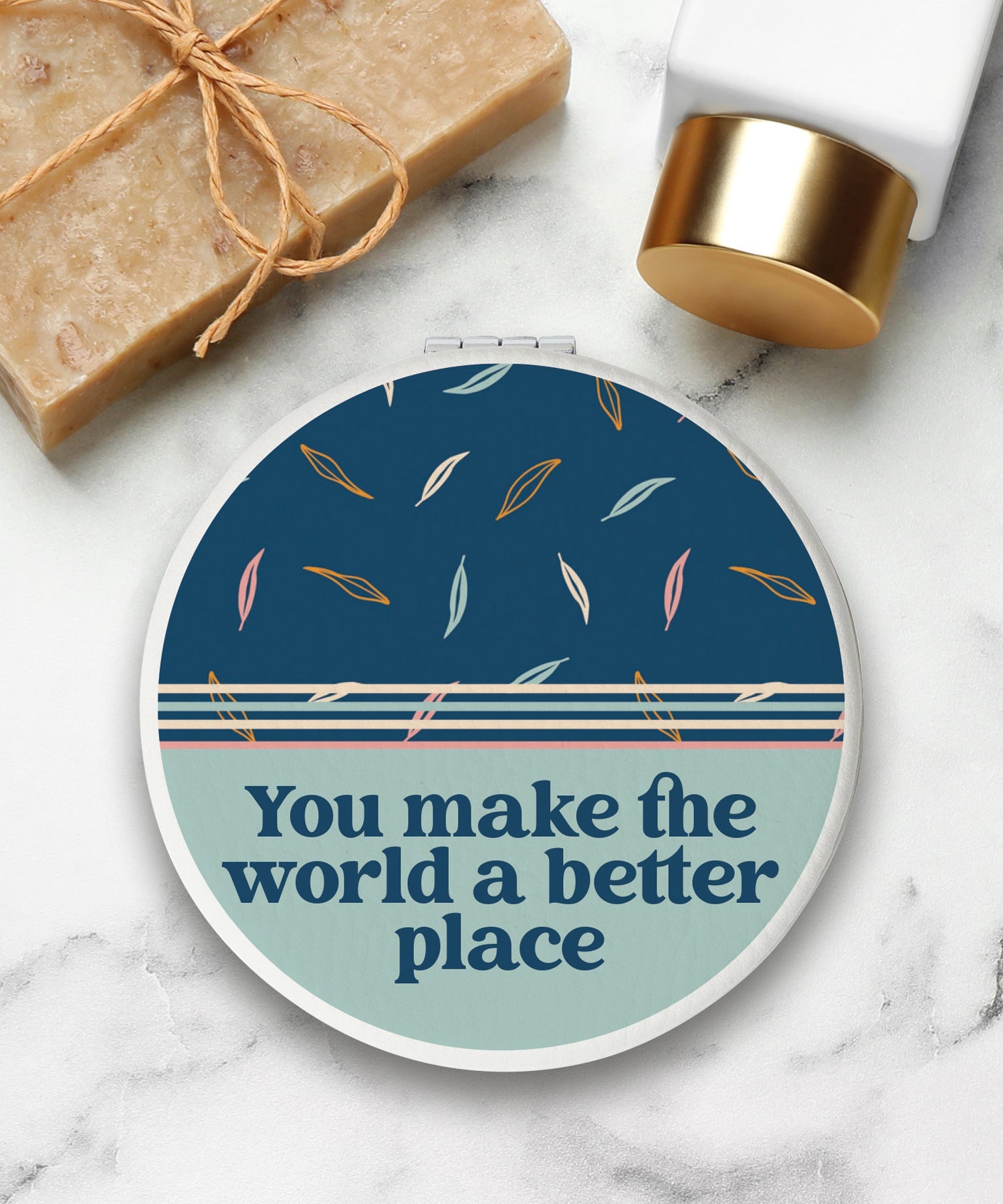 Round compact mirror with a leatherette cover, full-color “You Make the World a Better Place” design, 1X and 2X mirrors inside, and magnetic closure.